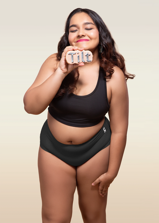 Woman wearing black sports bra and black mid-waist underwear, holding Tilt logo