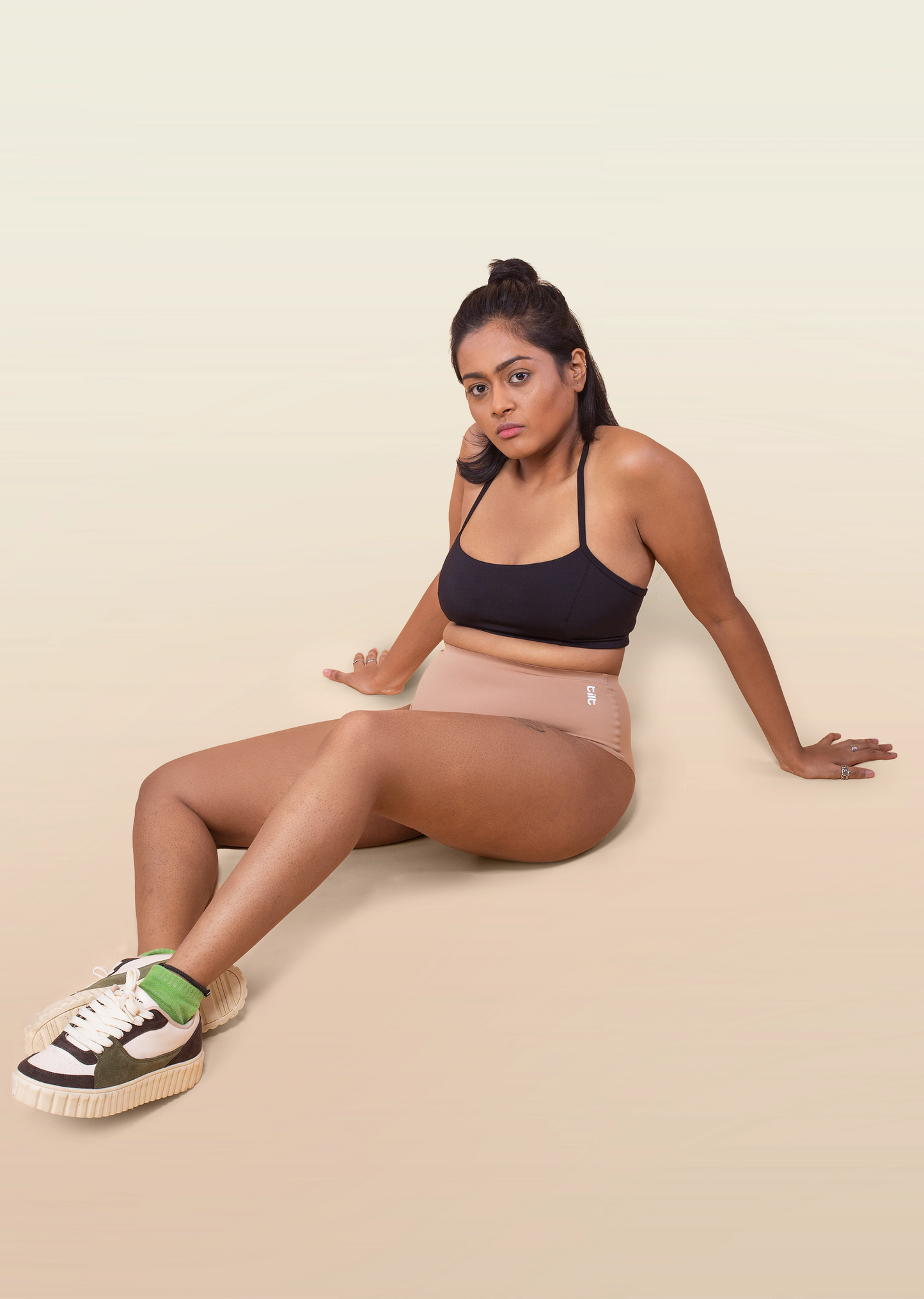 Woman sitting on a beige background wearing a black sports bra and beige underwear.