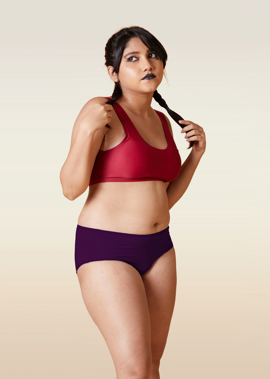 Woman wearing a red sports bra and violet mid-waist underwear against a beige background