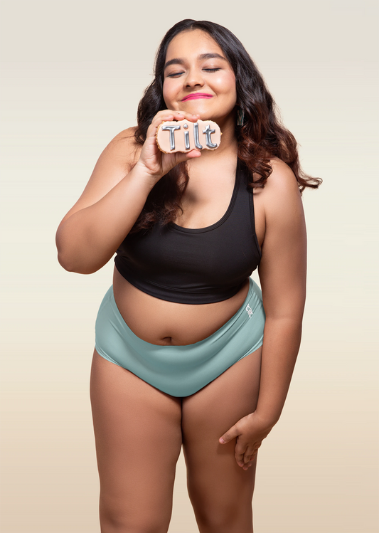 Woman wearing black sports bra and blue mid-waist seamless underwear holding a 'Tilt' sign.