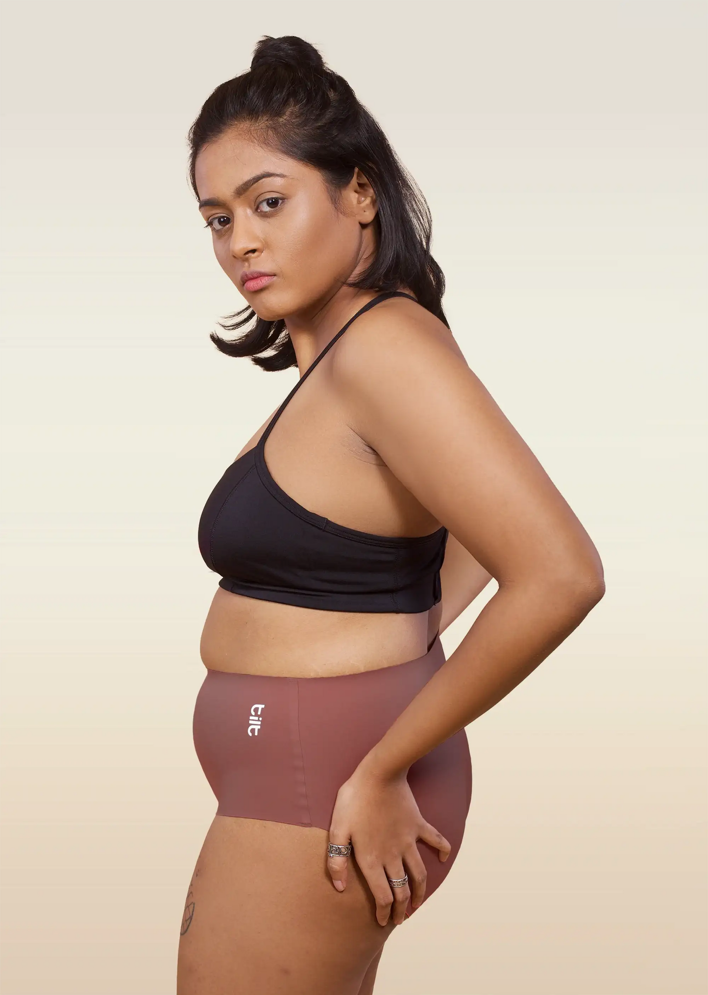 Woman wearing a black sports bra and pink mid-waist underwear on a beige background