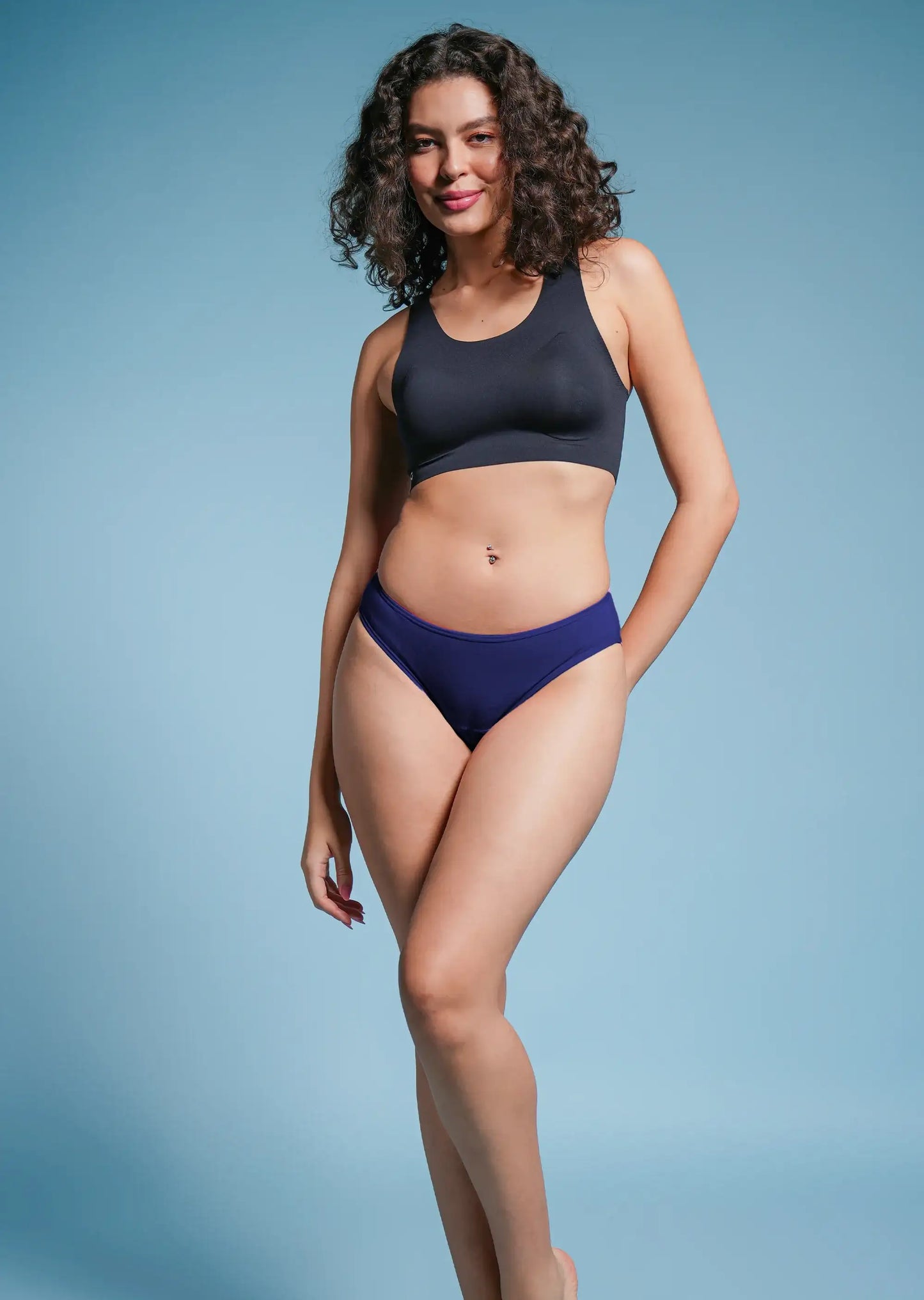 Woman wearing a black sports bra and blue low-waist bikini bottoms on a light blue background