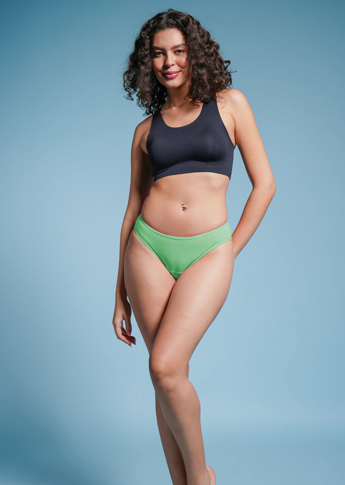 Woman wearing a black sports bra and green bikini bottoms on a blue background