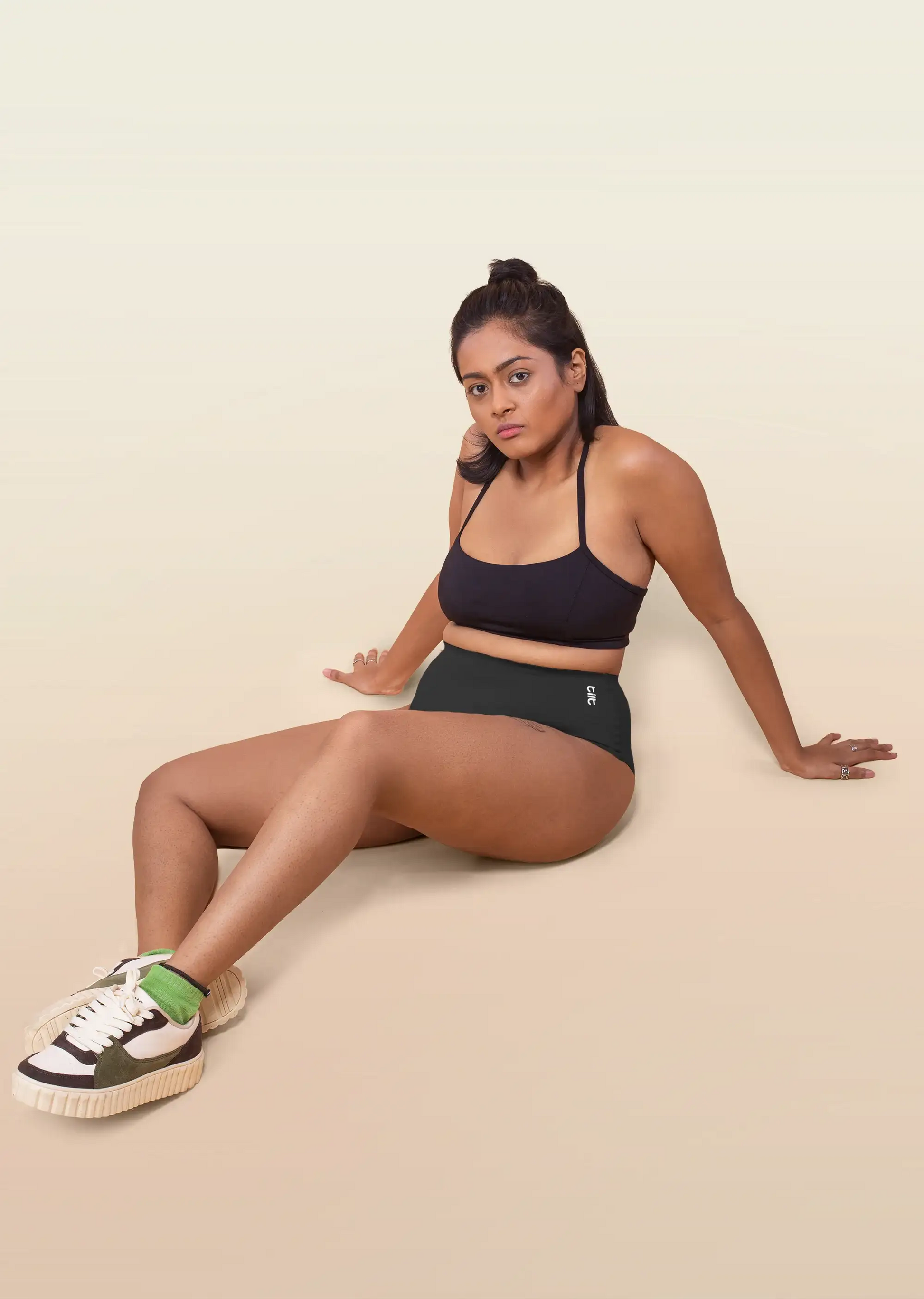 Woman sitting on a beige background wearing a black sports bra and underwear with sneakers.