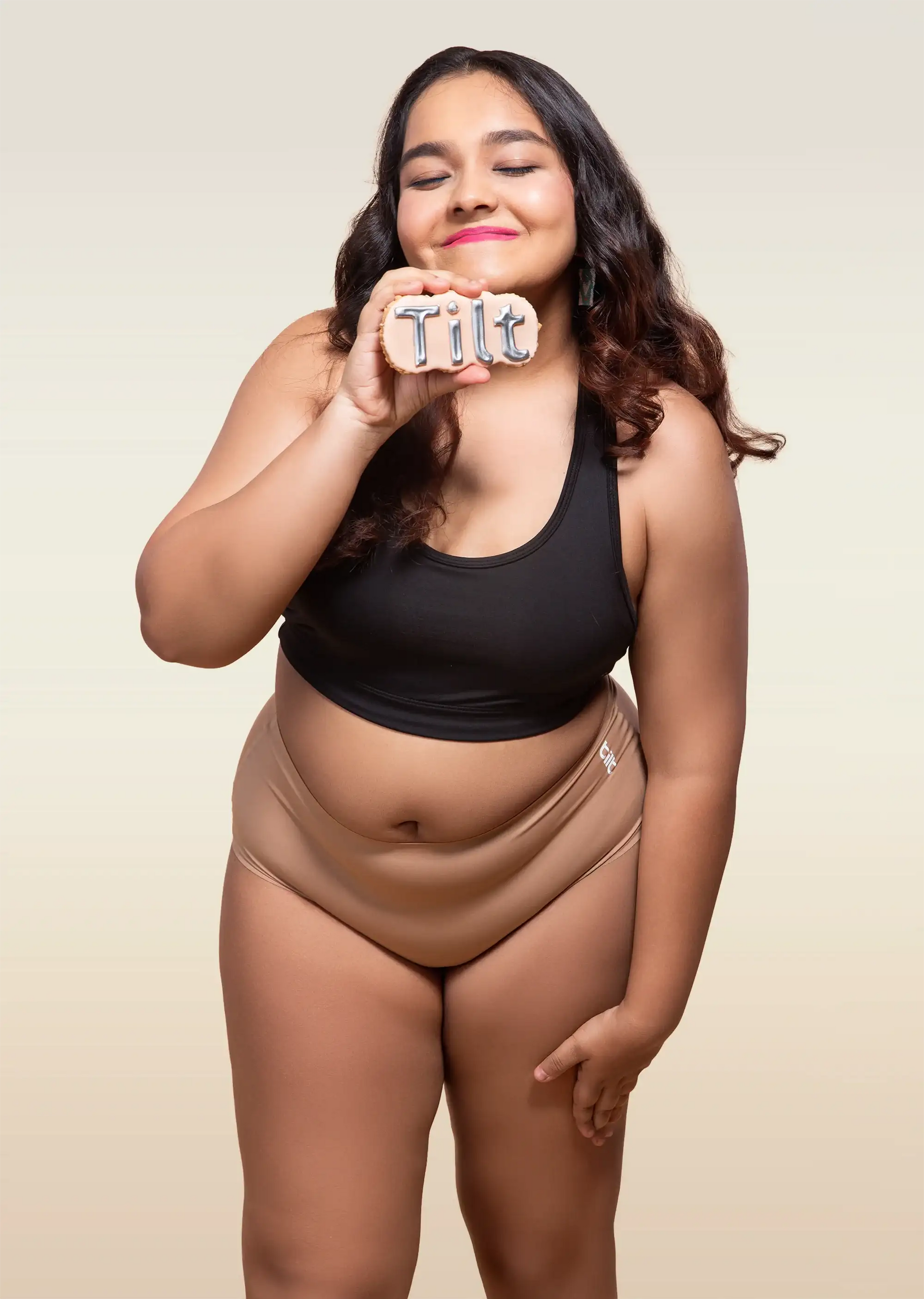 Woman wearing a black tank top and beige mid-waist underwear, holding a 'Tilt' sign.