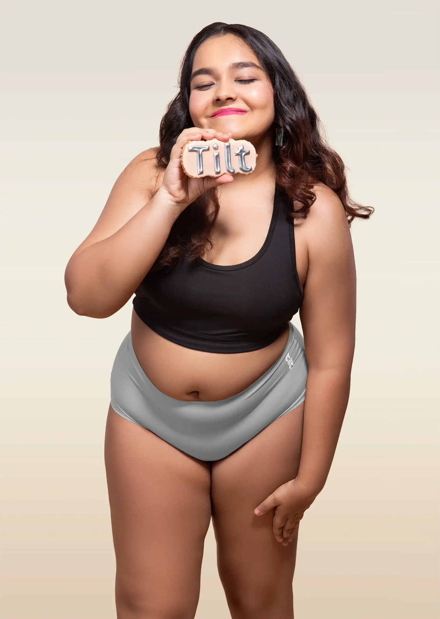 Woman wearing black sports bra and blue underwear, holding Tilt logo